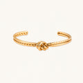 Gold cuff bracelet with twisted rope texture and decorative knot centerpiece on cream background.