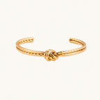 Gold cuff bracelet with twisted rope texture and decorative knot centerpiece on cream background.